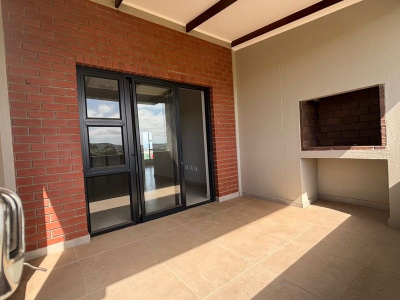 A modern covered balcony featuring a built-in braai, tiled flooring, and an exposed brick feature wall with large sliding glass doors.