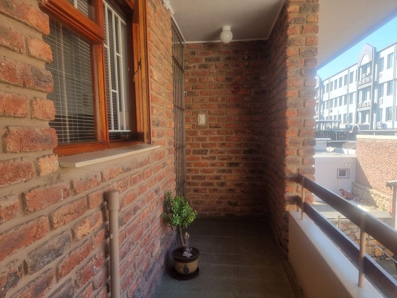 A covered balcony featuring exposed brick walls, tiled flooring, and a metal balustrade, offering an outdoor space with a view of the surrounding complex.