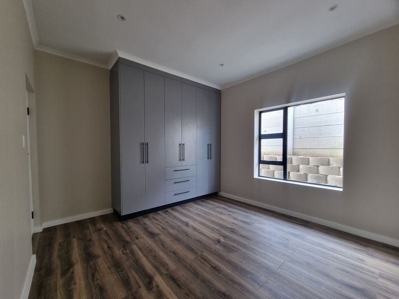 A clean, modern bedroom featuring neutral walls, durable laminate flooring, and ample built-in cupboard storage with a view of the exterior retaining wall.