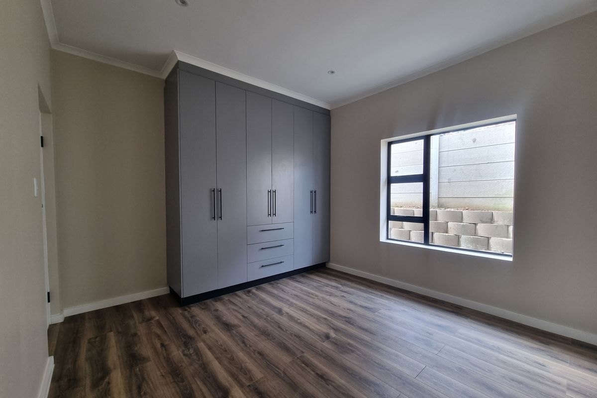 A clean, modern bedroom featuring neutral walls, durable laminate flooring, and ample built-in cupboard storage with a view of the exterior retaining wall.