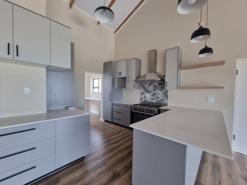 Modern kitchen featuring light grey cabinetry, quartz countertops, gas stove with extractor, and vaulted ceilings with exposed rafters, leading to a separate scullery area.