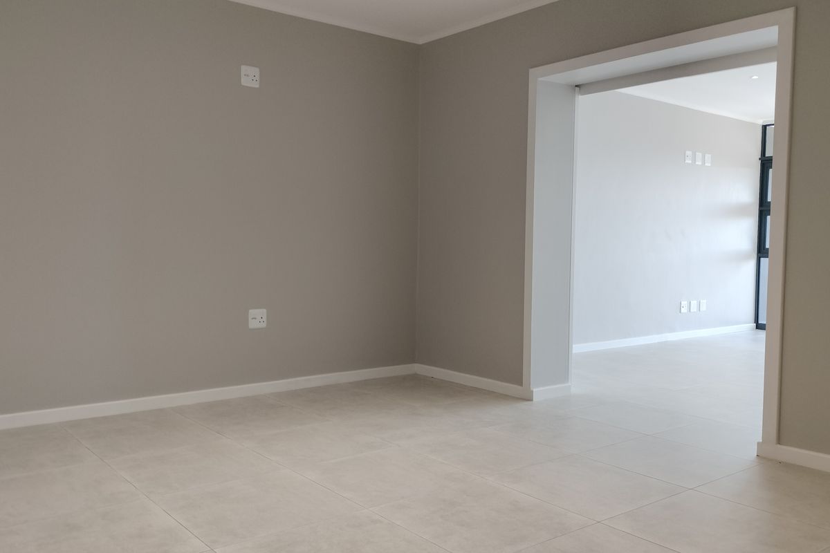 A bright, empty living area featuring neutral wall tones and modern tiled flooring, leading into an adjacent room through a wide doorway.
