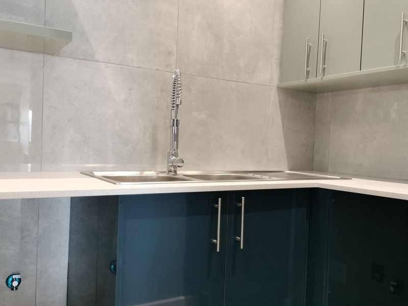 Modern kitchen area featuring sleek blue and sage green cabinetry, a quartz countertop, stainless steel sink, and contemporary grey tiled backsplash.