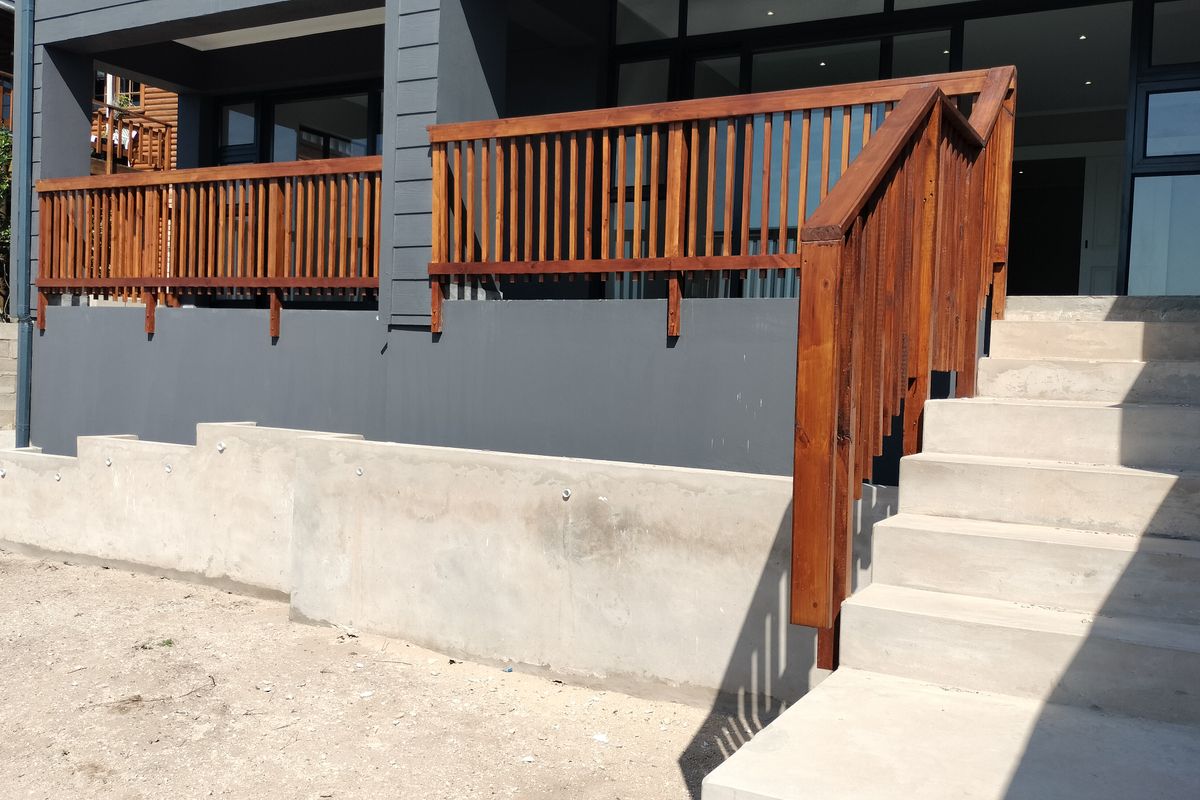 Rear exterior view featuring a timber balustrade deck, concrete staircase, and unfinished retaining wall leading to the main living area sliding doors.