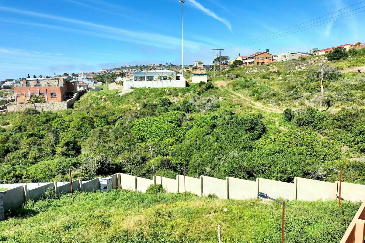 An elevated view of a sloped residential plot featuring natural vegetation, perimeter walling, and surrounding neighborhood infrastructure in the Garden Route District.