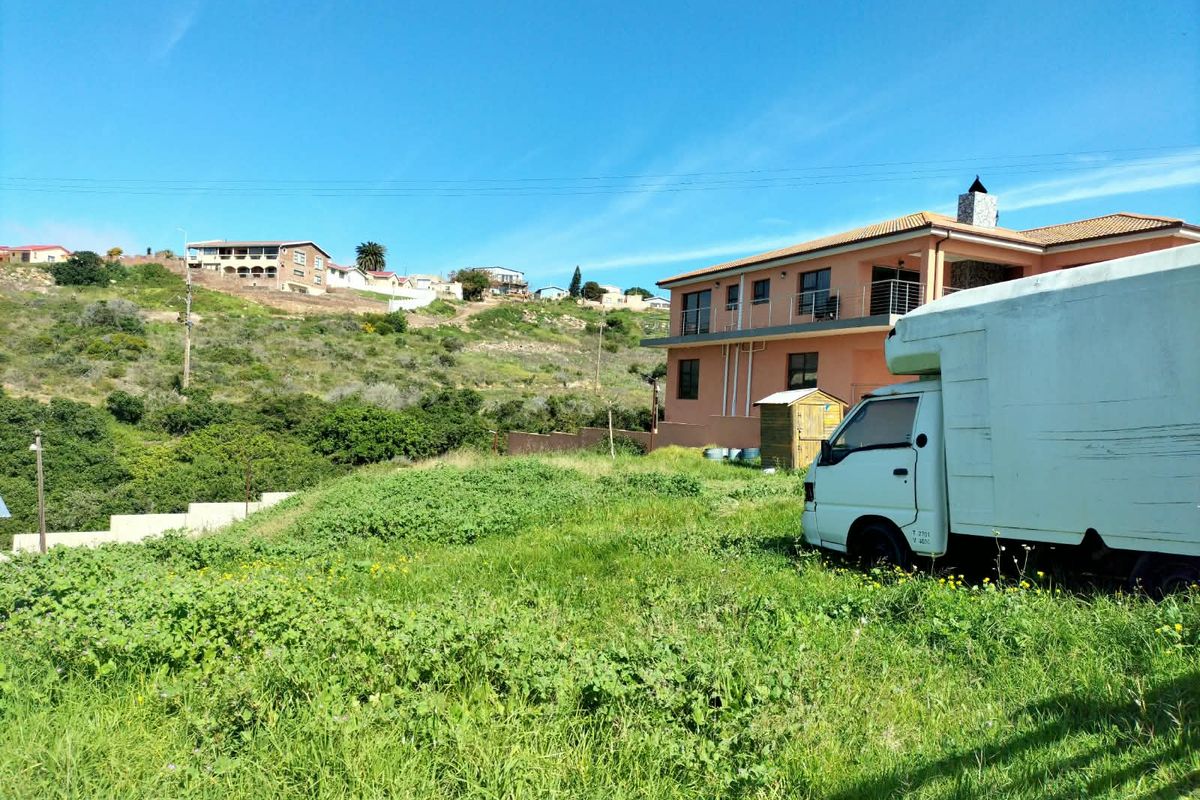 A view of a sloped residential plot with a multi-story building in the background, featuring natural vegetation and a retaining wall structure.