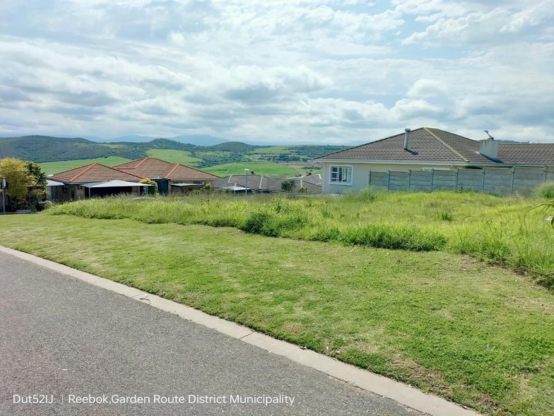 A vacant residential plot situated in a developed neighborhood, featuring a gentle slope, grass cover, and scenic views of the surrounding hills.