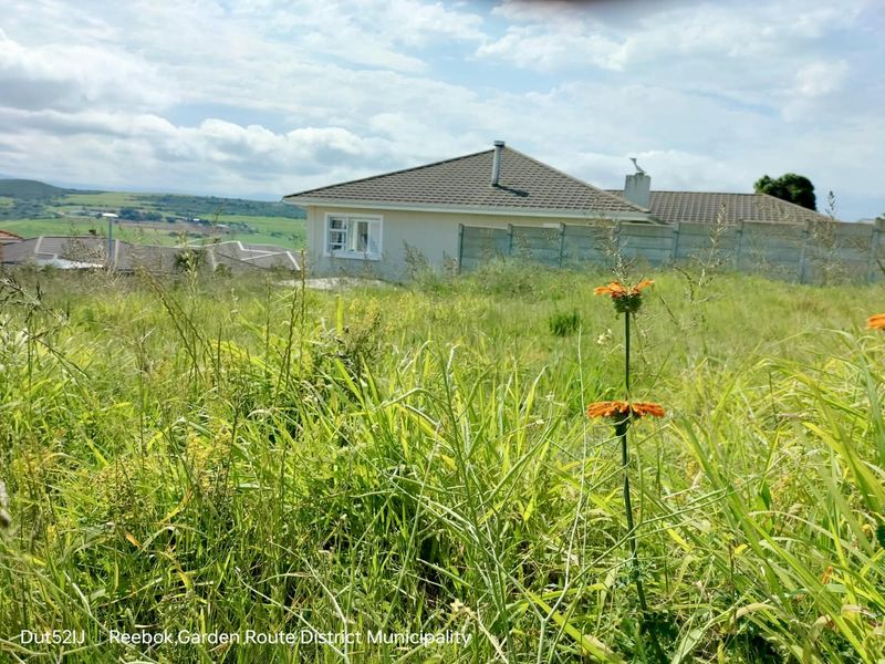 A view of a vacant residential plot featuring overgrown grass with a neighboring house and boundary wall visible in the background.