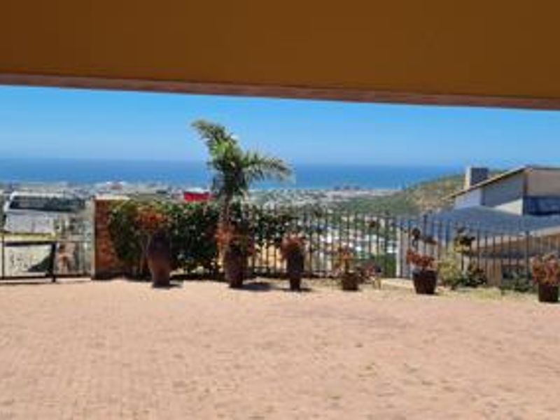 A covered patio area featuring paved flooring and metal fencing, offering an expansive elevated view of the surrounding landscape and distant coastline.
