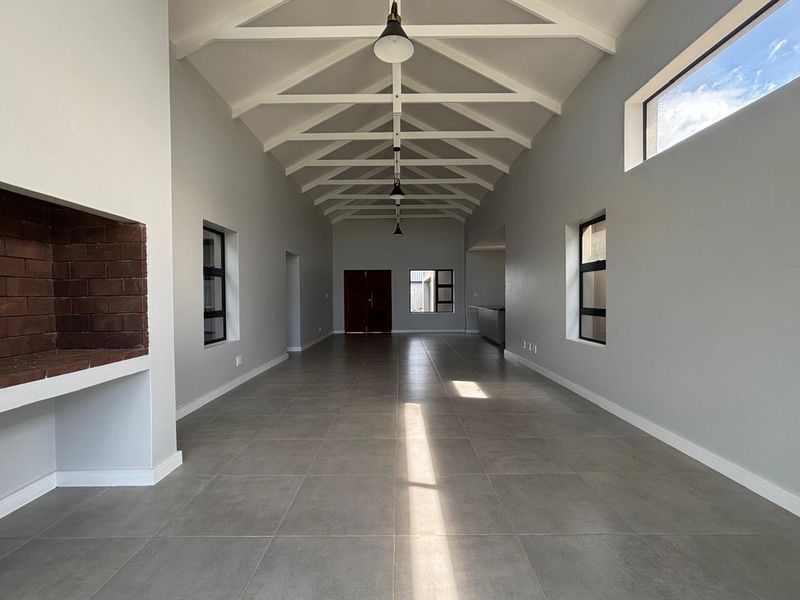 Spacious open-plan living area featuring high vaulted ceilings with exposed trusses, large format floor tiles, and a built-in brick fireplace.