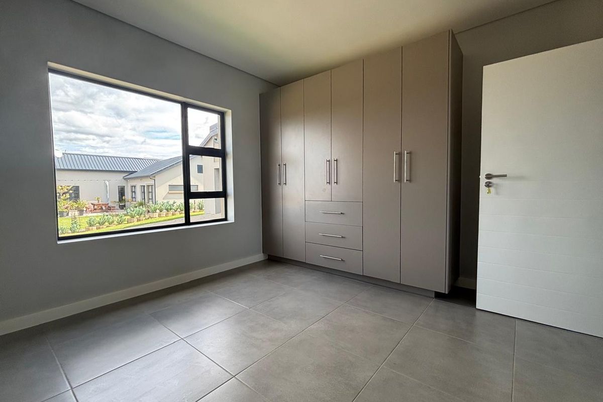 A bright, modern bedroom featuring neutral grey floor tiles, ample built-in cupboard storage, and a large window overlooking the garden.