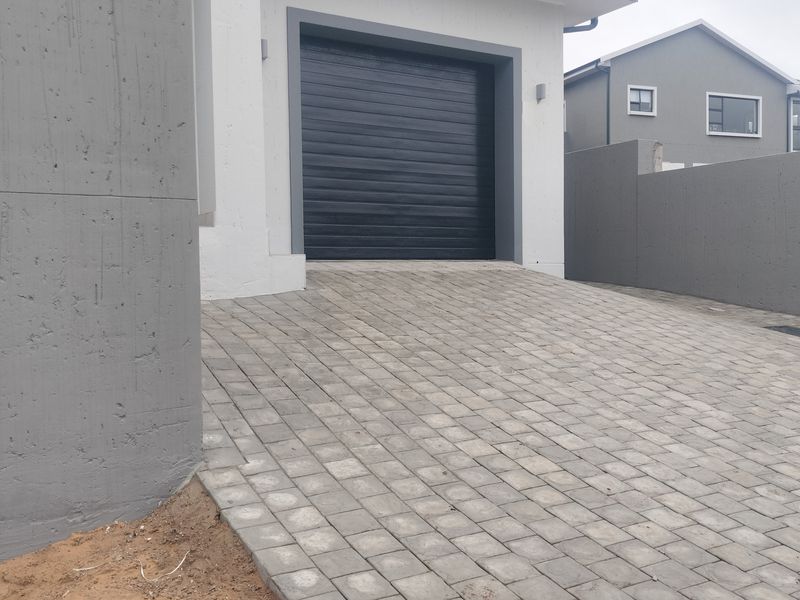 A modern residential exterior featuring a paved driveway leading to a single sectional garage door, enclosed by concrete boundary walls.