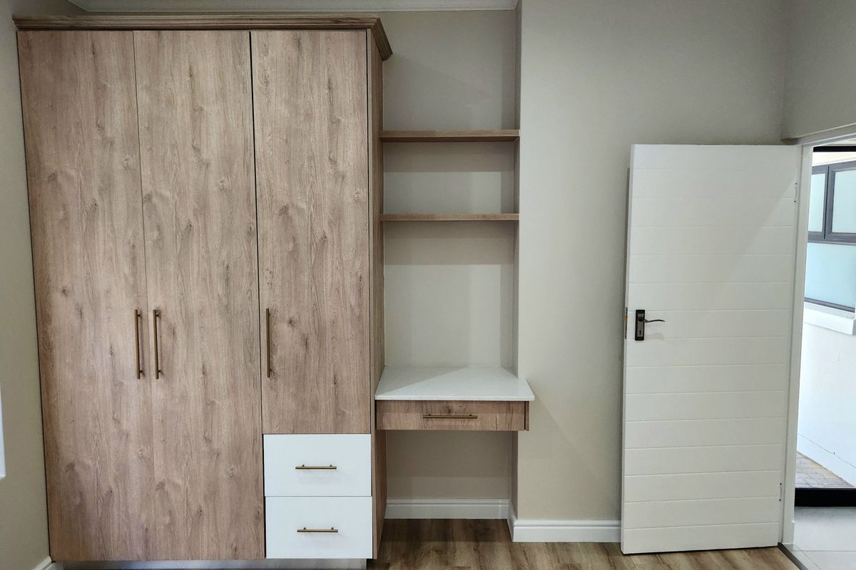 A modern bedroom featuring a built-in wood-grain wardrobe with an integrated desk and shelving unit, complemented by laminate flooring.