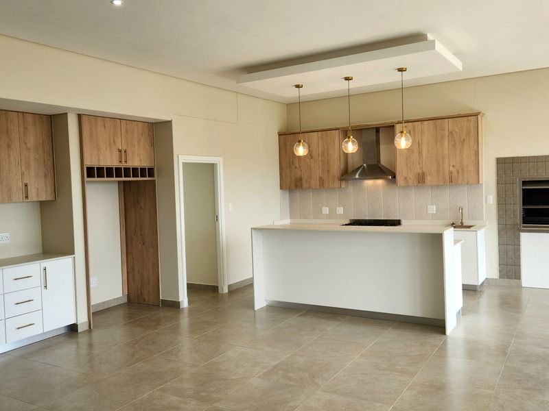 Spacious open-plan kitchen featuring modern wood-finish cabinetry, a central island with stone countertops, and a built-in indoor braai area.