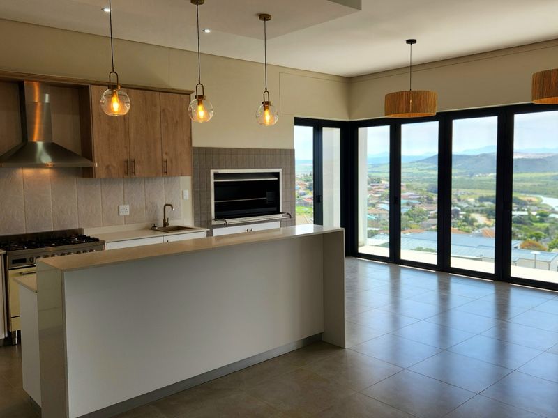 Modern open-plan kitchen and living area featuring a built-in braai, sleek cabinetry, gas hob, and expansive stacking doors with panoramic views.