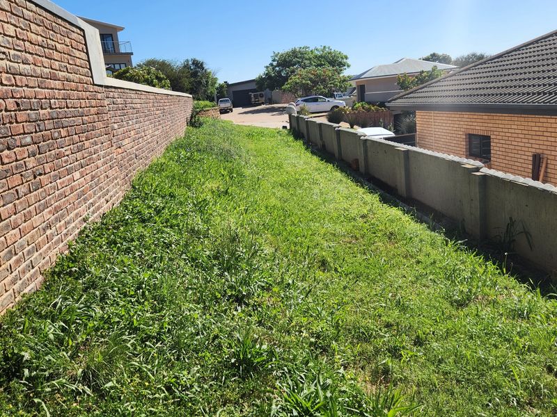 A narrow strip of residential land enclosed by a brick boundary wall and a precast concrete wall, featuring overgrown grass.