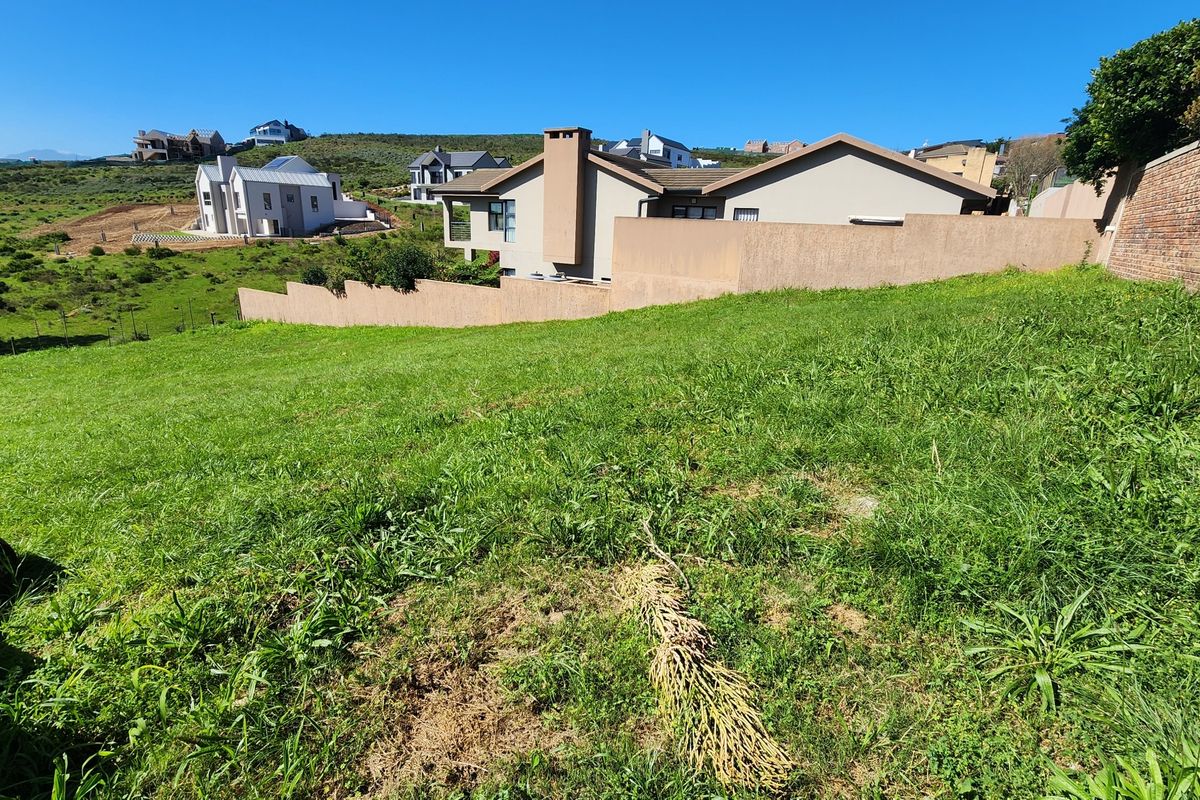 A view of a vacant residential plot featuring a grassy slope, boundary walls, and surrounding neighborhood homes.