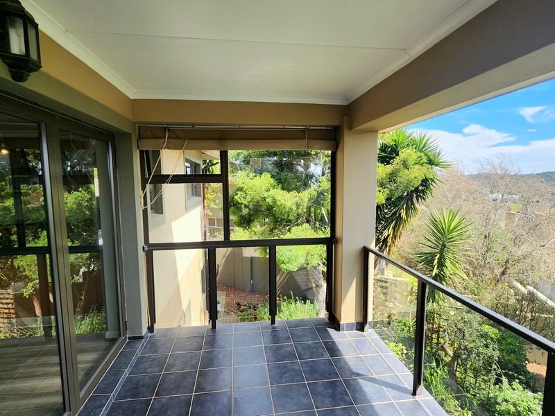 A covered balcony featuring tiled flooring, a modern glass balustrade, and sliding door access, offering elevated views of the surrounding garden and landscape.