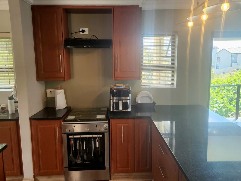 A functional kitchen featuring warm-toned wooden cabinetry, dark granite countertops, a gas stove with extractor fan, and direct access to an outdoor patio area.