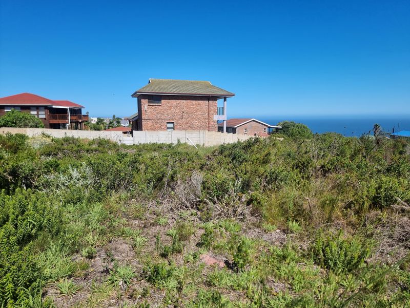 A vacant residential plot featuring coastal vegetation, enclosed by a precast concrete boundary wall with views of neighboring properties and the ocean.