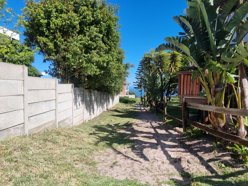 A sandy pathway bordered by a precast concrete boundary wall and timber fencing, featuring mature trees and coastal vegetation.