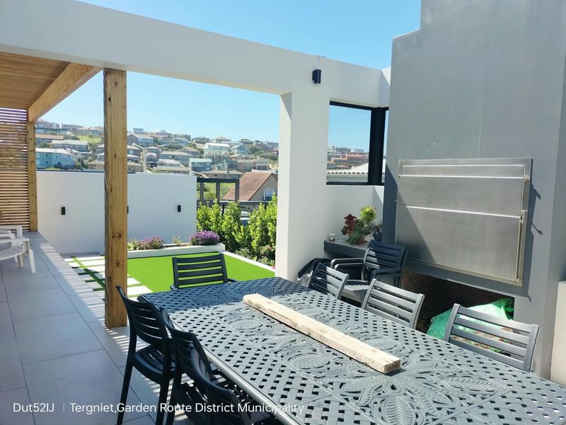 A modern covered patio featuring a built-in braai, tiled flooring, and a small landscaped garden area with artificial lawn, offering pleasant neighborhood views.