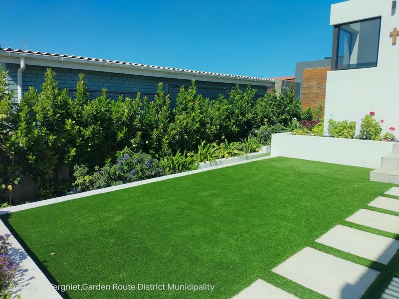 A well-maintained garden area featuring artificial turf, concrete stepping stones, and a raised planter box against a boundary wall.