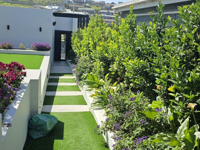 A well-maintained garden featuring artificial lawn, paved pathways, and raised concrete planters with established greenery and vibrant flowering shrubs.