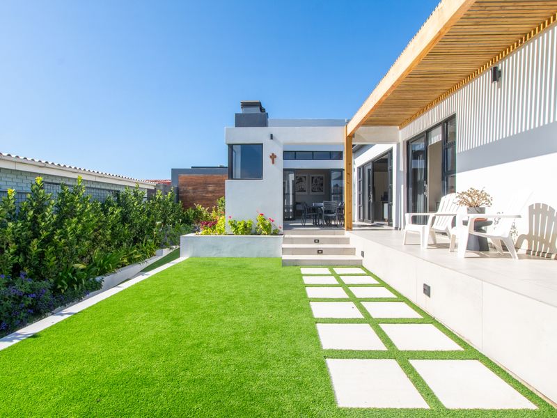 Modern rear exterior featuring a tiled patio with timber pergola, built-in fireplace, and a well-maintained artificial lawn with stepped garden beds.