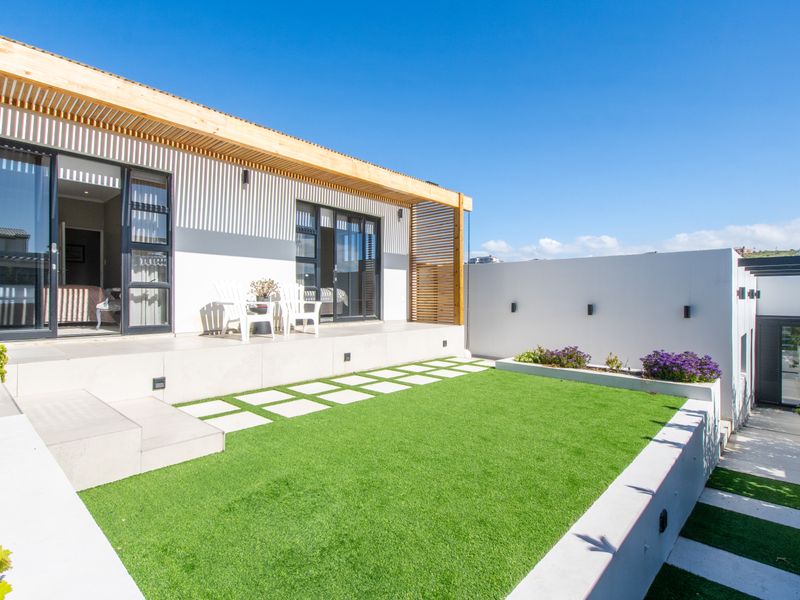 A modern rear exterior featuring a timber pergola, sliding glass doors, artificial lawn, and tiered concrete landscaping.