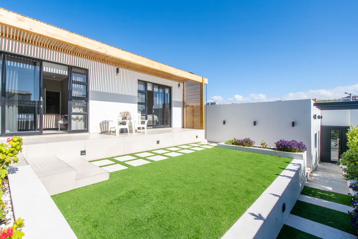 A modern rear exterior featuring a timber pergola, sliding glass doors, artificial lawn, and tiered concrete landscaping.