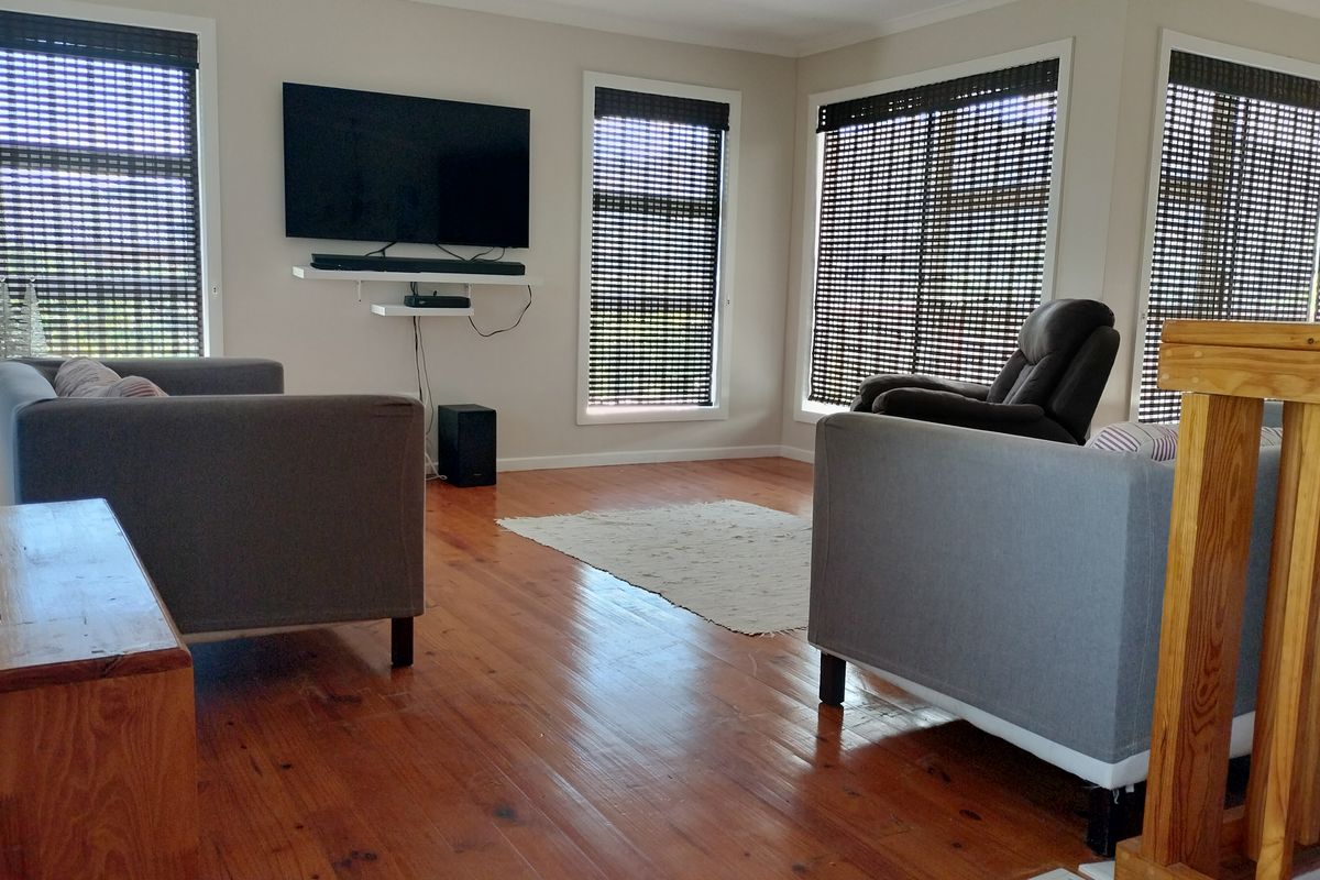 A bright living room featuring polished timber flooring, a timber balustrade, and multiple windows fitted with dark roller blinds.