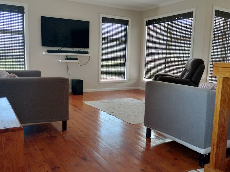A bright living room featuring polished timber flooring, a timber balustrade, and multiple windows fitted with dark roller blinds.