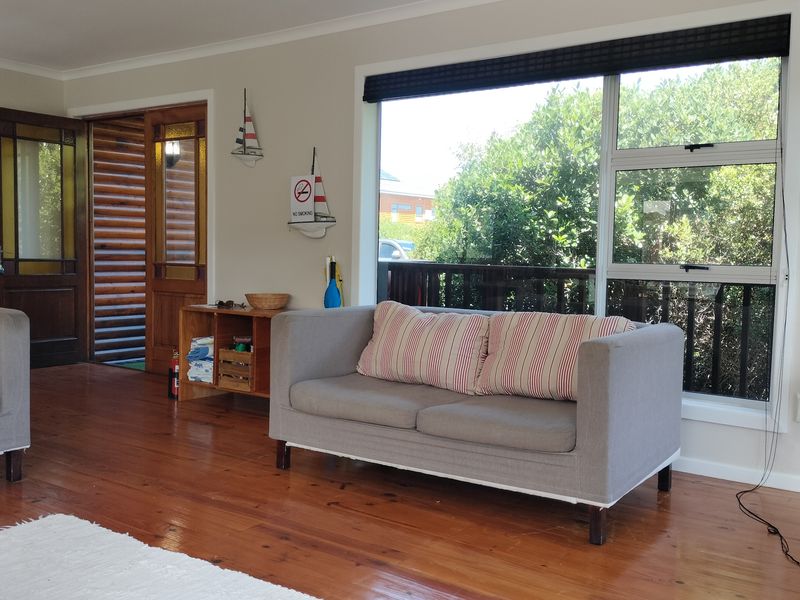A bright living room featuring polished timber flooring, a large window with natural light, and a classic timber entrance door.