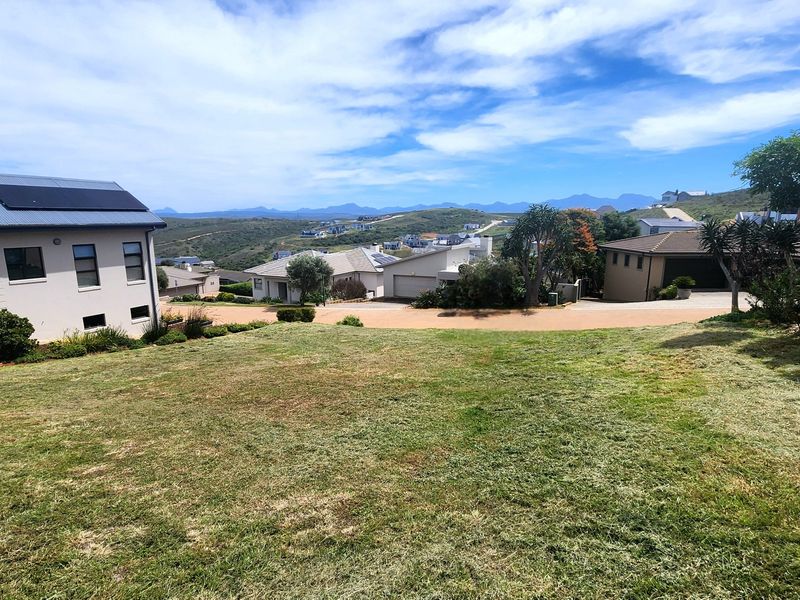 A view of a sloped residential plot featuring a maintained grass lawn with surrounding neighborhood homes and distant mountain scenery.