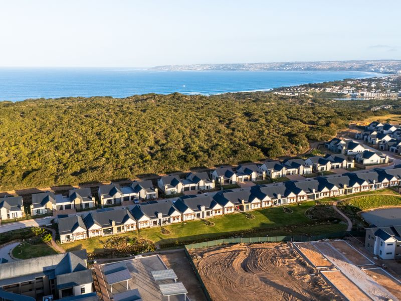 An aerial view of a modern residential cottage estate situated near the coastline, featuring uniform architectural styles and well-maintained communal green spaces.