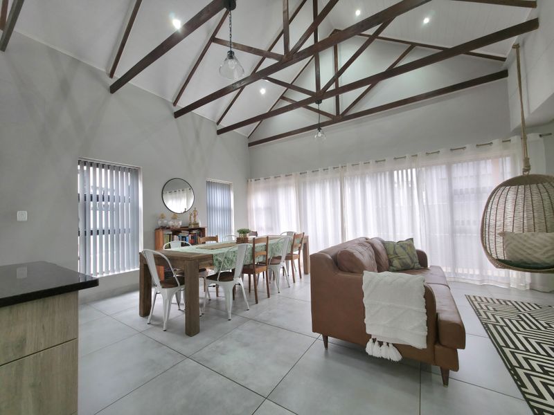 Spacious open-plan living and dining area featuring high vaulted ceilings with exposed timber trusses, large windows with vertical blinds, and modern tiled flooring.