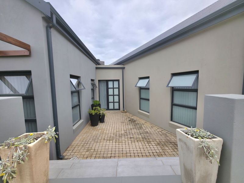 A private paved courtyard area connecting two wings of the house, featuring modern grey and beige exterior walls and multiple awning windows.