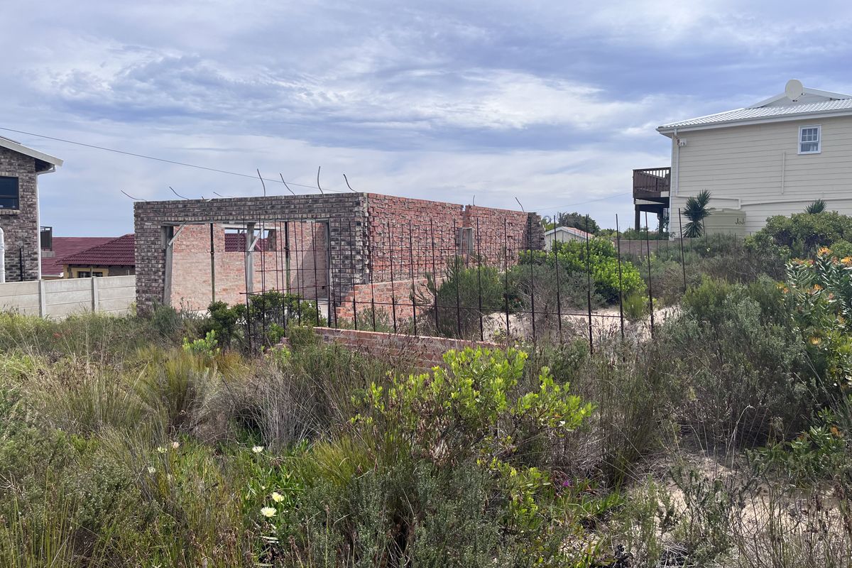 An unfinished brick structure on a vacant residential plot, showing exposed masonry and reinforcing steel bars, indicating a project currently under construction.