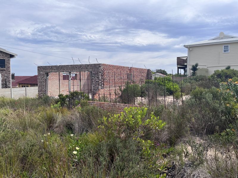 An unfinished brick structure on a vacant residential plot, showing exposed masonry and reinforcing steel bars, indicating a project currently under construction.