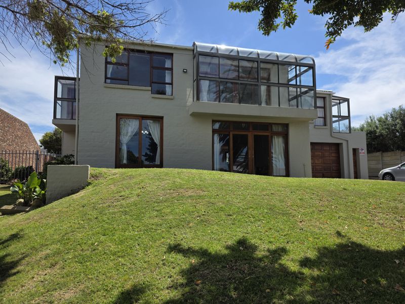 Rear view of a multi-story house featuring a prominent glass-enclosed balcony, sliding doors, and a large sloped lawn area.