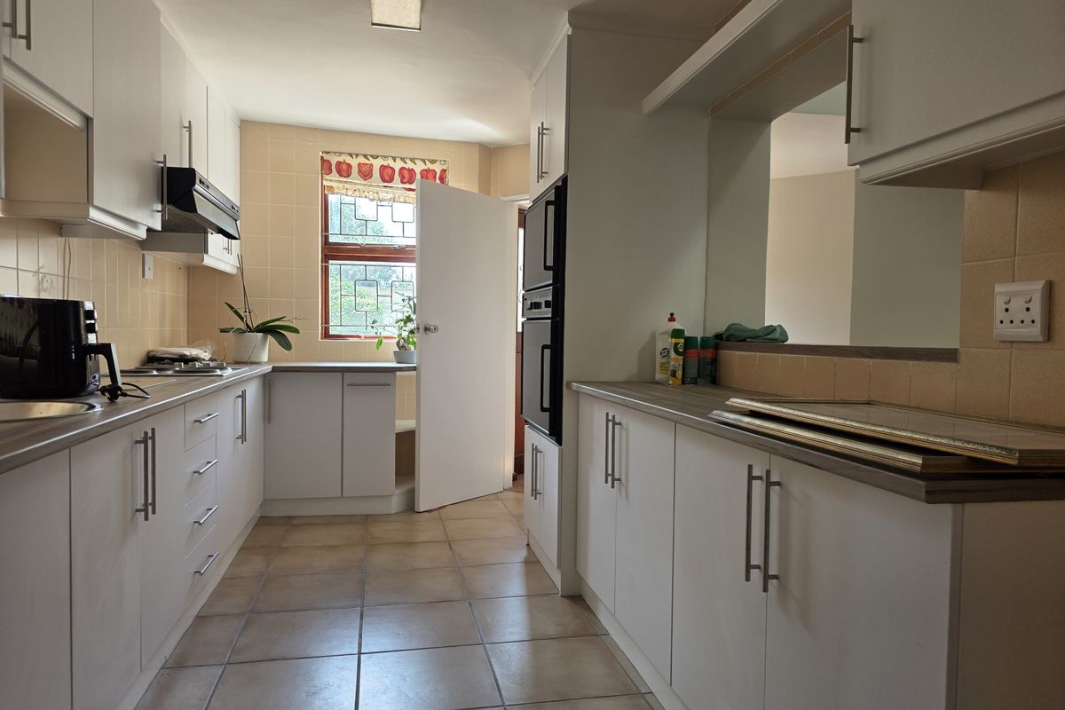A functional galley-style kitchen featuring white cabinetry, laminate countertops, built-in double oven, and tiled flooring with a window providing natural light.