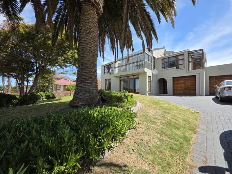 Front elevation of a multi-story house featuring a paved driveway, double garage, and a prominent glass-enclosed upper balcony set within a landscaped garden.