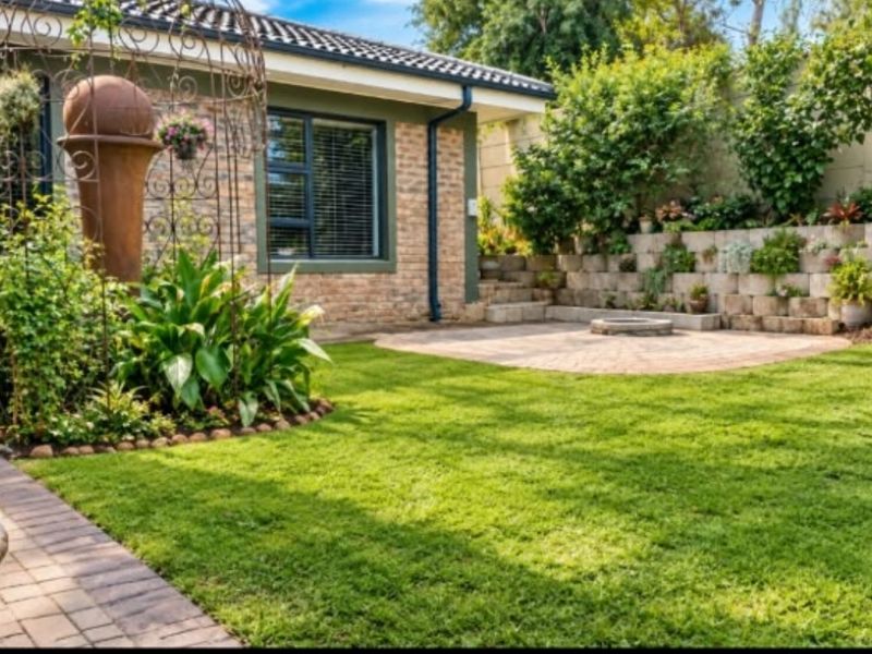 A well-maintained rear garden featuring a paved patio area, lush lawn, and tiered retaining wall against a brick-clad house exterior.