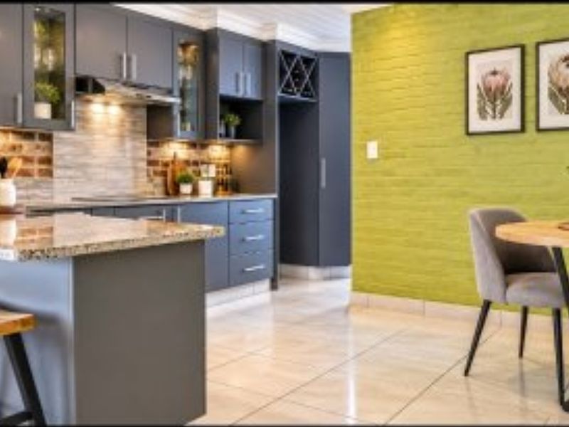 Modern open-plan kitchen featuring dark cabinetry, granite island, and exposed brick accents, seamlessly flowing into a bright dining area with tiled flooring.