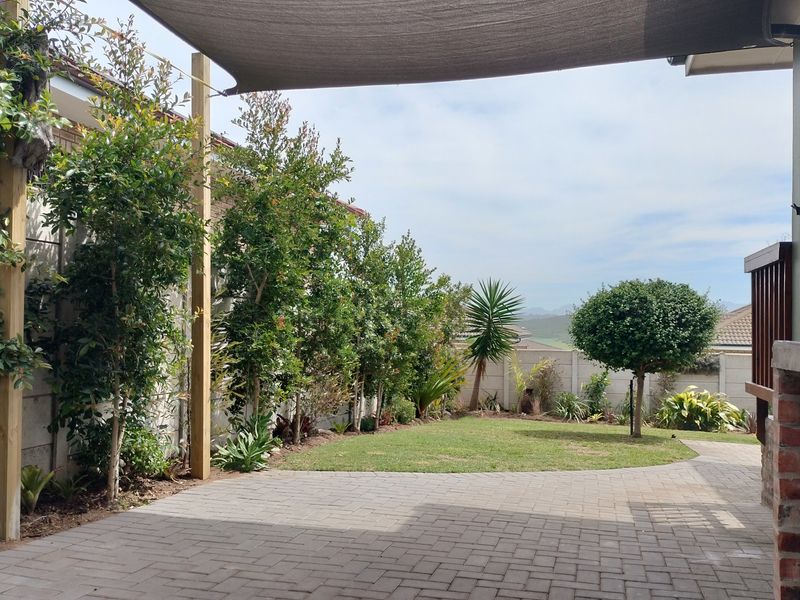 A well-maintained paved patio area featuring a shade sail, overlooking a neat garden with established trees and boundary walling.