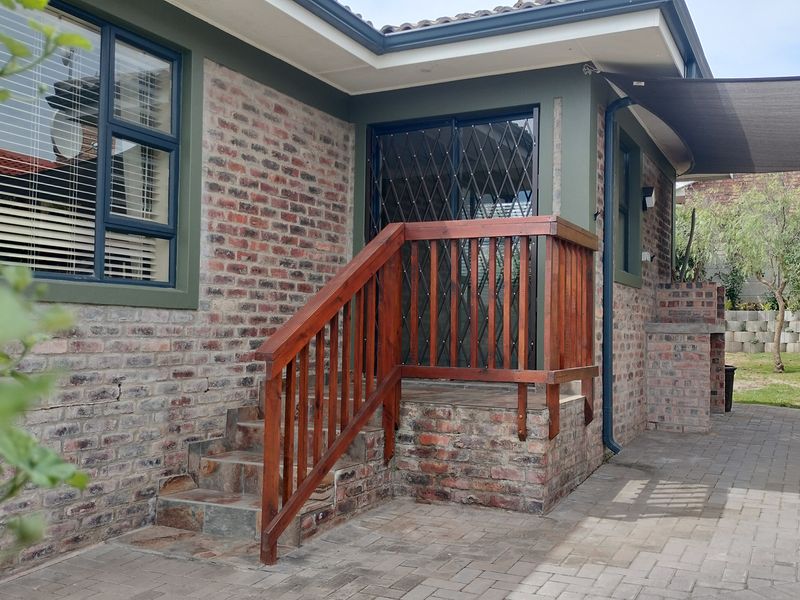 Rear exterior view featuring a paved patio, built-in braai, timber staircase with railing, and brick facade with modern aluminium window frames.