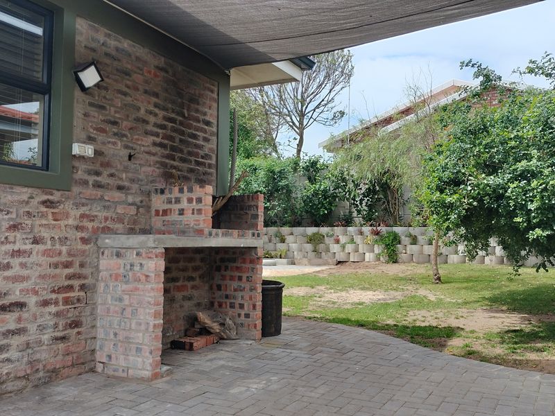 Covered patio area featuring a brick built-in braai, paved flooring, and a shade sail, overlooking a landscaped garden with a retaining wall.