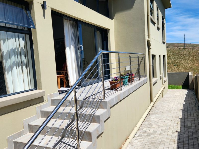 An elevated tiled patio area featuring a modern stainless steel balustrade, accessible via sliding glass doors, with an adjacent paved walkway and boundary wall.