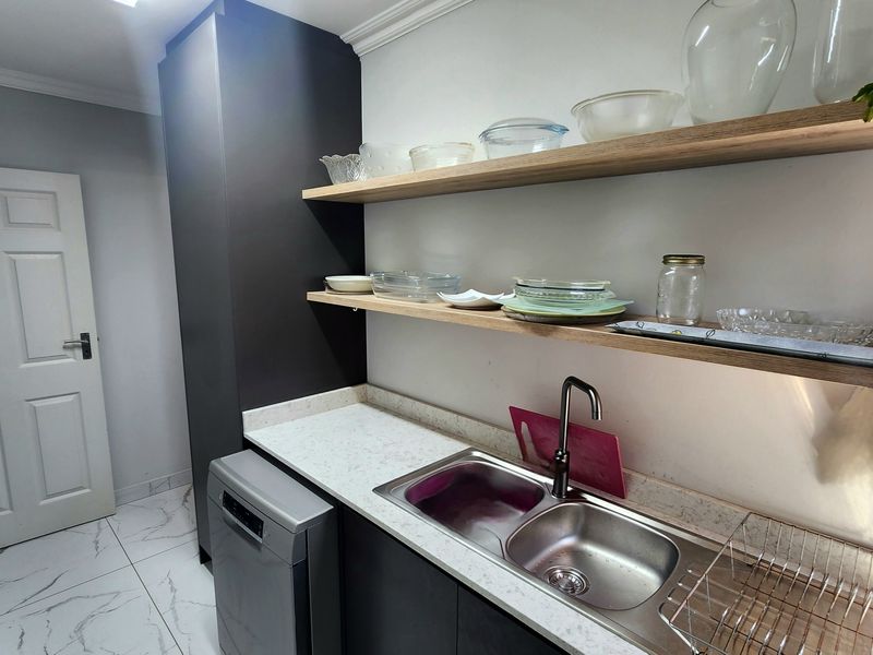 A modern scullery featuring stone countertops, a double bowl stainless steel sink, floating timber shelving, and sleek dark cabinetry with tiled flooring.